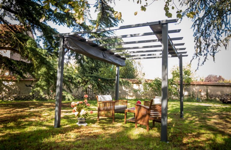 Pergola installée dans un jardin, aménagée avec des fauteuils en bois pour créer un espace détente à l'ombre. Illustration d'un agencement extérieur avec pergola par Bricomarché Cosne.