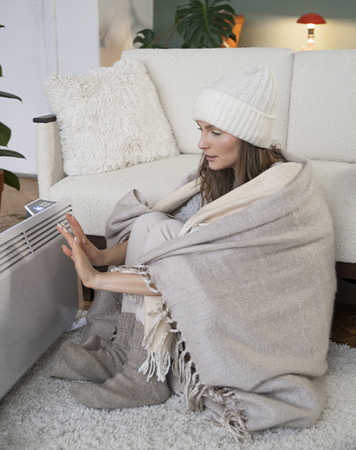 Femme emmitouflée dans une couverture et portant un bonnet, assise près d'un radiateur pour se réchauffer dans un salon confortable. Illustration de solutions de chauffage intérieur proposées par Bricomarché Cosne.