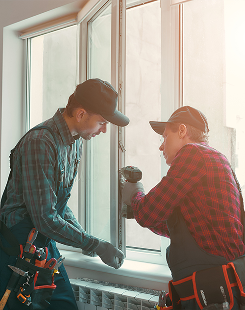 Deux artisans en chemise à carreaux installant une fenêtre avec un outil électrique, illustrant le service de pose de fenêtres proposé par Bricomarché Cosne.