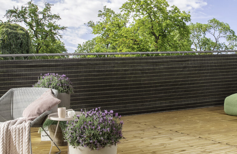 Terrasse en bois aménagée avec des pots de lavande, un fauteuil confortable avec coussin et couverture, et une table basse. La terrasse est entourée d'un brise-vue en matériau tissé pour plus d'intimité. Exemple d'aménagement extérieur proposé par Bricomarché Cosne.