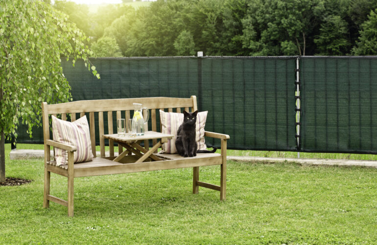 Jardin aménagé avec un banc en bois, décoré de coussins à rayures, et une petite table avec une carafe et des verres, entouré de verdure et protégé par une clôture verte. Exemple d'agencement extérieur par Bricomarché Cosne.