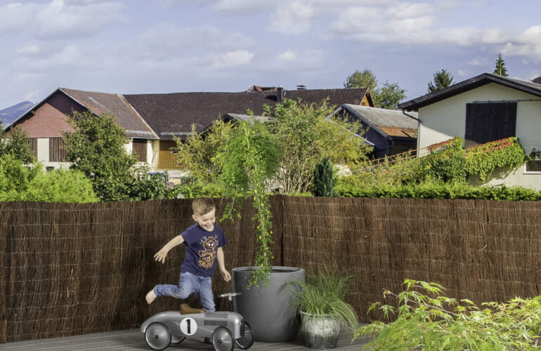 Petit garçon jouant sur une terrasse aménagée avec un véhicule jouet, entouré de plantes en pots et d'une clôture en brande de bruyère. Illustration d'un agencement extérieur par Bricomarché Cosne.