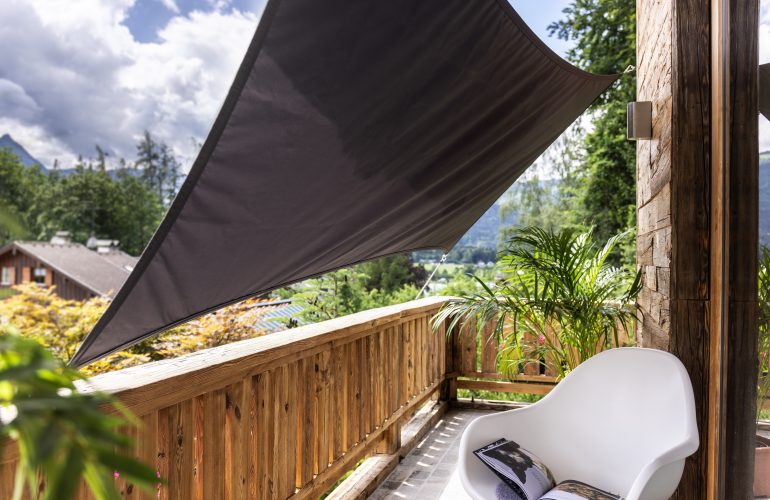 Balcon en bois avec une voile d'ombrage triangulaire grise offrant un espace de détente avec une chaise blanche et des plantes vertes. Agencement extérieur par Bricomarché Cosne.